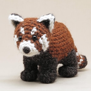 May include: A crocheted red panda toy. The soft toy is made with brown yarn for the body, black for the legs, and white and brown for the face. It has black eyes and a small black nose. The tail is striped with brown and white.
