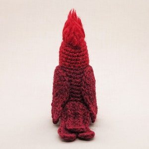 May include: A crocheted cardinal bird plush toy with a red and burgundy ombre color scheme. The bird is facing away from the camera, showing its back and tail feathers.