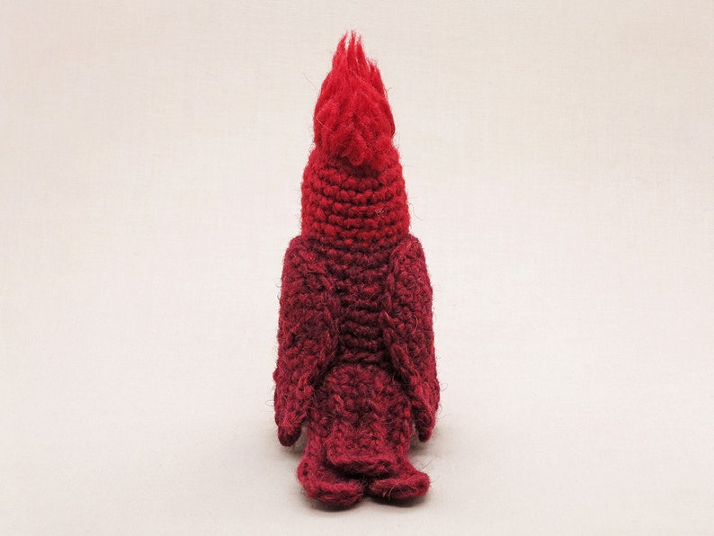 May include: A crocheted cardinal bird plush toy with a red and burgundy ombre color scheme. The bird is facing away from the camera, showing its back and tail feathers.