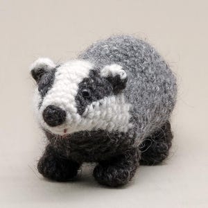 May include: A small, handcrafted crochet badger figurine featuring a textured grey back, dark charcoal legs, and a white face with black eye patches. The animal has a round black nose, small rounded ears, and two dark, glossy bead eyes.