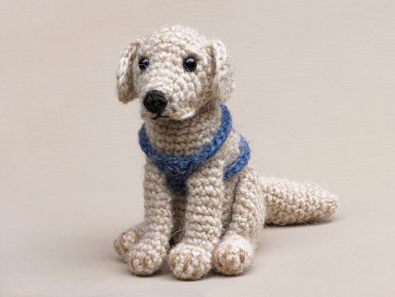 May include: A crocheted stuffed dog toy with a light brown body and blue collar. The dog is sitting with its legs crossed and its tail curled behind it.