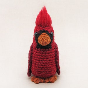 May include: A crocheted cardinal bird toy with a red body, black eyes, and an orange beak. The bird has a fluffy red crest on its head.
