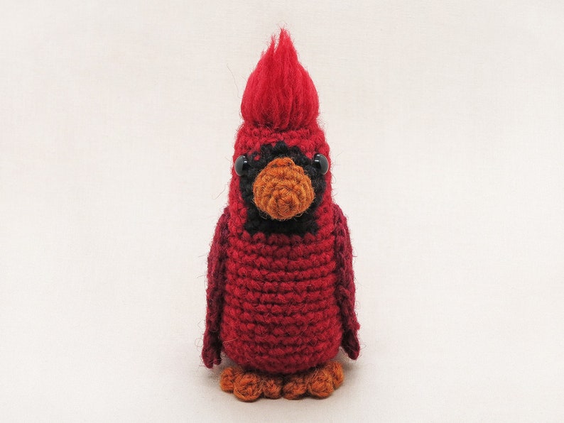 May include: A crocheted cardinal bird toy with a red body, black eyes, and an orange beak. The bird has a fluffy red crest on its head.