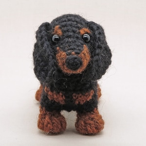 May include: A black and brown crocheted dachshund toy with black eyes. The toy is standing on a white background.