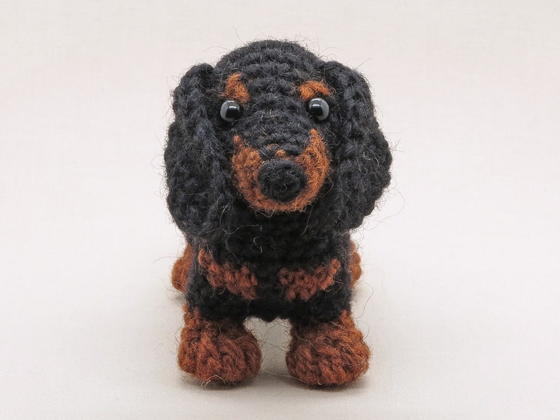 May include: A black and brown crocheted dachshund toy with black eyes. The toy is standing on a white background.
