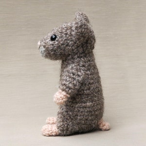 May include: A gray crocheted mouse toy with black eyes and pink feet. The mouse is standing on a light brown surface.