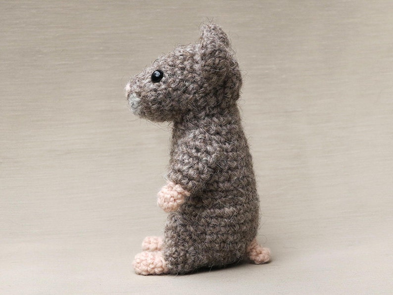 May include: A gray crocheted mouse toy with black eyes and pink feet. The mouse is standing on a light brown surface.