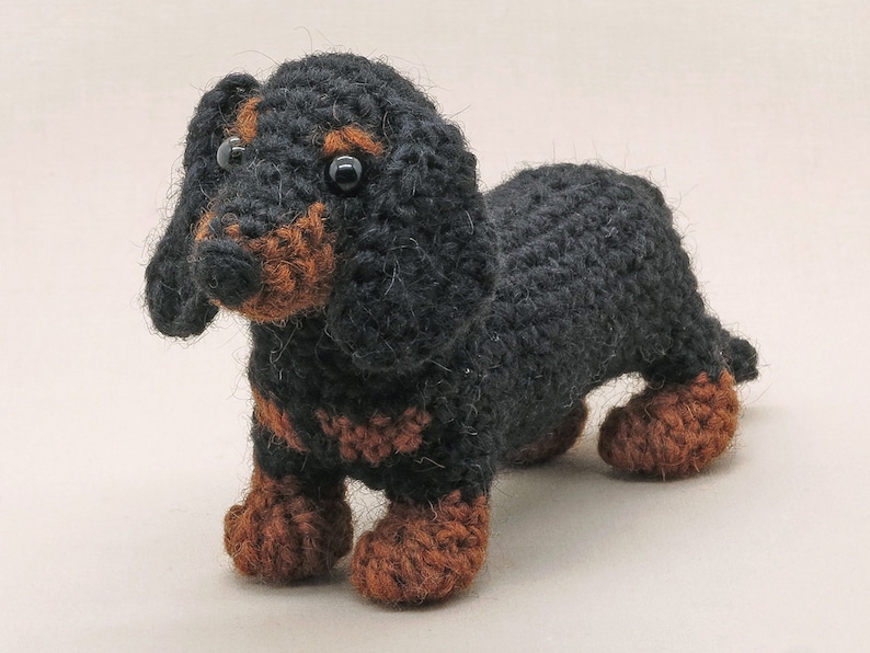 May include: A black and brown crocheted dachshund toy. The toy has black fur and brown accents on its legs and face.