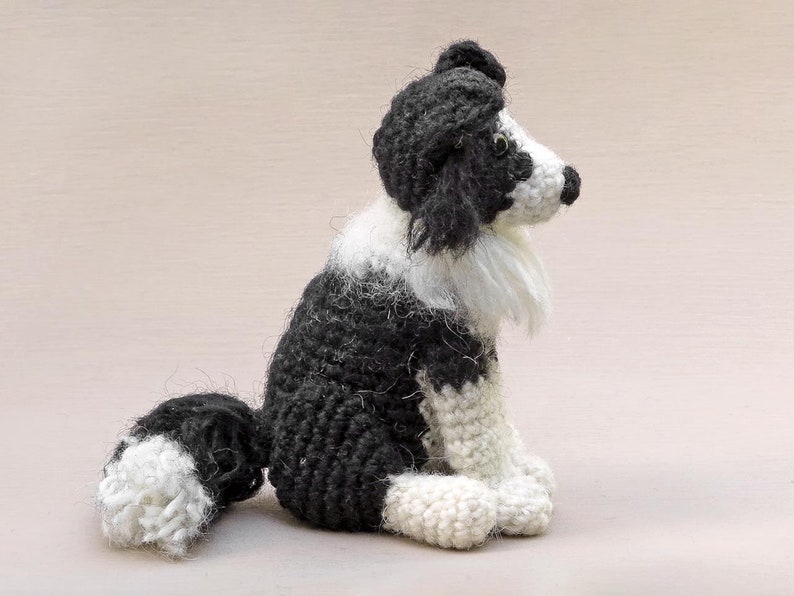 May include: A crocheted black and white border collie dog sitting with its head turned to the side. The dog has a fluffy white tail and a white patch on its chest.