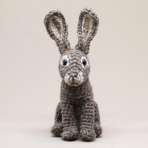 May include: A handmade, gray crocheted hare with long, upright ears and blue eyes. The amigurumi-style rabbit is sitting upright, showcasing its detailed texture and craftsmanship. This soft sculpture is a charming decorative item.