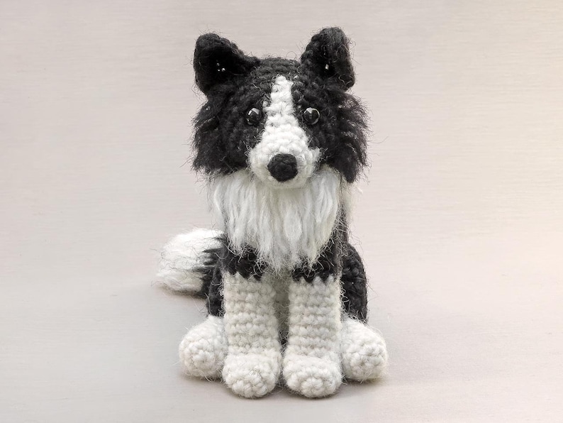 May include: A crocheted black and white border collie dog sitting with its paws crossed. The dog has a white face and chest with black fur on its back and tail.