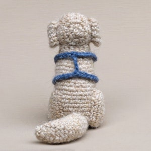 May include: A crocheted dog toy with a blue harness. The dog is sitting with its tail curled up behind it.