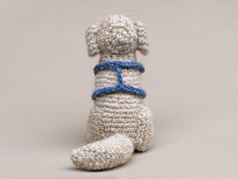 May include: A crocheted dog toy with a blue harness. The dog is sitting with its tail curled up behind it.