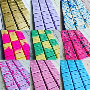 May include: Assortment of wax melts in various colors and patterns. The wax melts are arranged in rows of six, with each row featuring a different color and pattern. The colors include gold, purple, green, pink, blue, and white. The patterns include solid colors, stripes, and splatters.