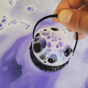 May include: A round, white bath bomb with purple detailing and a black handle, creating a bubbly, foamy effect. The bath bomb is held above purple-coloured bath water, with bubbles floating on the surface.