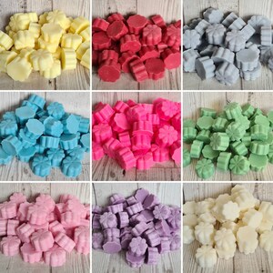 May include: Assortment of wax melts in various colors and shapes, including white, yellow, red, pink, blue, purple, green, and gray. The wax melts are shaped like flowers and are arranged in piles on a wooden surface.