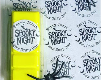 May include: A yellow wax snap bar in a clear plastic container. The label reads "Berry Cocoa Spooky Night Wax Snap Bar" in a spooky font. Decorative black spiders and matching labels are scattered around the bar.