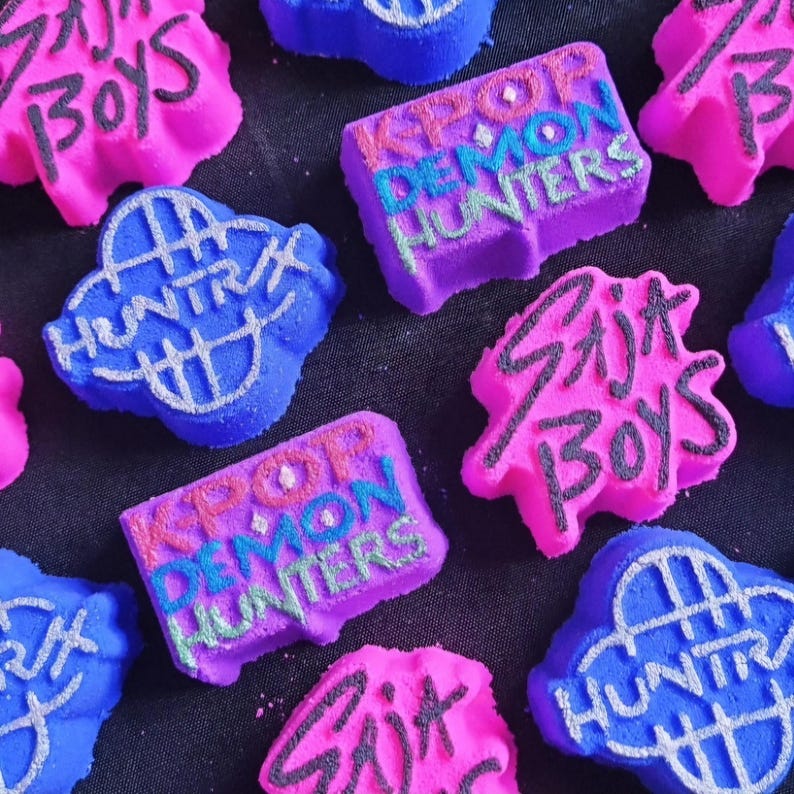 May include: A selection of vibrantly coloured bath bombs. The bath bombs are in pink, purple, and blue, with text that reads "KPOP DEMON HUNTERS", "Skit Boys", and "HUNTR".