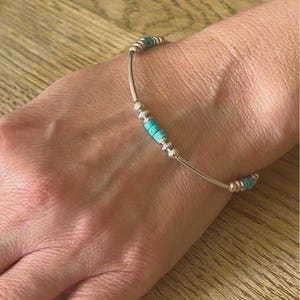 May include: A silver bracelet featuring turquoise beads and silver detailing. The bracelet has a curved design and is worn on a wrist. The turquoise beads are cylindrical and spaced along the silver band.