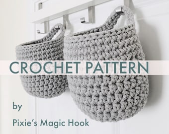 May include: Two gray crocheted hanging baskets with a white background. The text "CROCHET PATTERN" is displayed in a banner above the baskets. The text "by Pixie's Magic Hook" is displayed below the baskets.