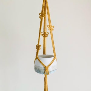 Op de afbeelding: Een macram&eacute; plantenhanger met een witte keramische pot. De hanger is gemaakt van geel koord en heeft een knoopmotief. De pot heeft een blauwe en witte glazuur.