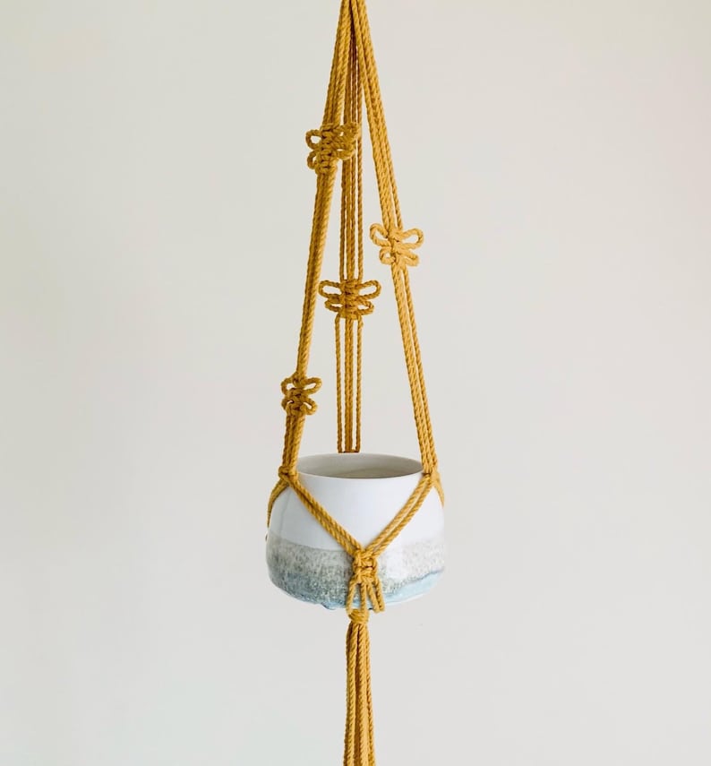 Op de afbeelding: Een macram&eacute; plantenhanger met een witte keramische pot. De hanger is gemaakt van geel koord en heeft een knoopmotief. De pot heeft een blauwe en witte glazuur.