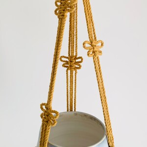 Op de afbeelding: Een macram&eacute; plantenhanger gemaakt van geel katoenen touw met vier knopen. De hanger houdt een lichtblauwe keramische pot.