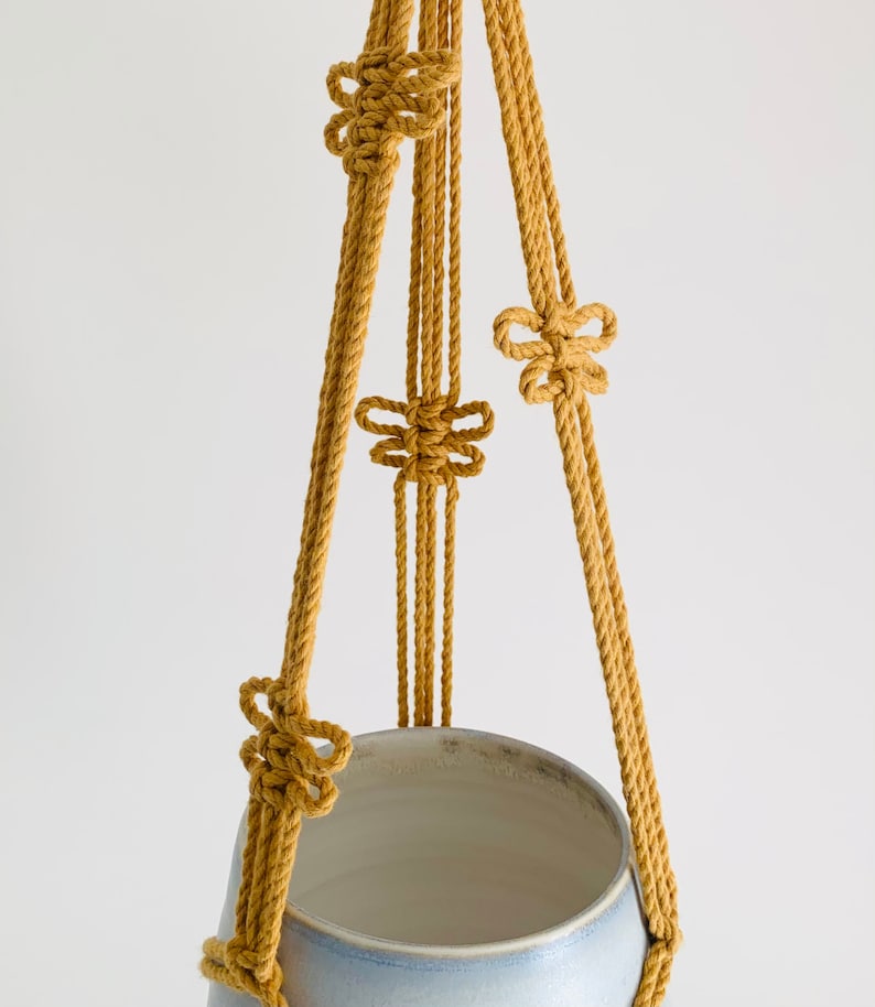 Op de afbeelding: Een macram&eacute; plantenhanger gemaakt van geel katoenen touw met vier knopen. De hanger houdt een lichtblauwe keramische pot.