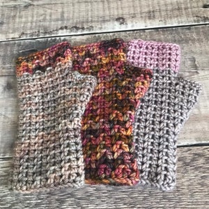 May include: Four crocheted fingerless gloves in different colors and patterns. The gloves are made with a variety of yarns, including a speckled gray, a multi-colored yarn with shades of brown, orange, and pink, and a solid gray yarn.