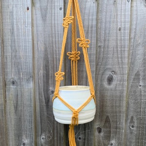 Op de afbeelding: Een macram&eacute; plantenhanger gemaakt van mosterdgele touw, met een witte keramische pot die eraan hangt.