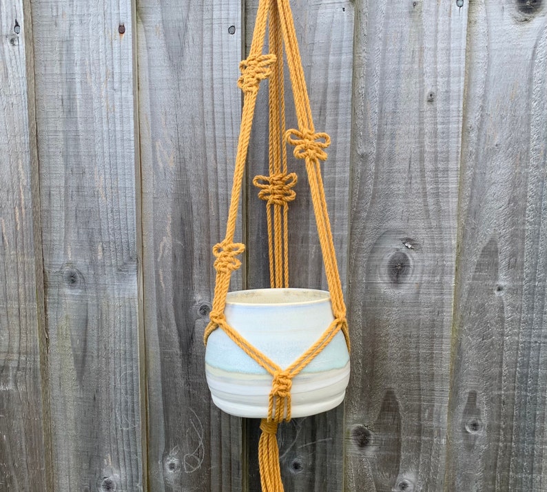 Op de afbeelding: Een macram&eacute; plantenhanger gemaakt van mosterdgele touw, met een witte keramische pot die eraan hangt.