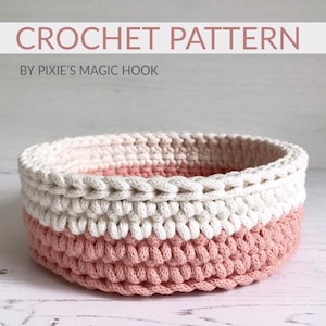 Pode incluir: Uma cesta de crochê redonda, com um design de duas cores. A base é rosa claro, a seção do meio é branca e a borda superior é branca. O texto "CROCHET PATTERN" é visível. Ideal para armazenamento ou decoração.
