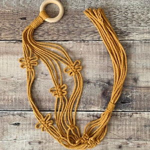 Op de afbeelding: Een macram&eacute; plantenhanger met een houten ring aan de bovenkant en vijf geknoopte bloemmotieven. De hanger is gemaakt van naturel gekleurd koord.