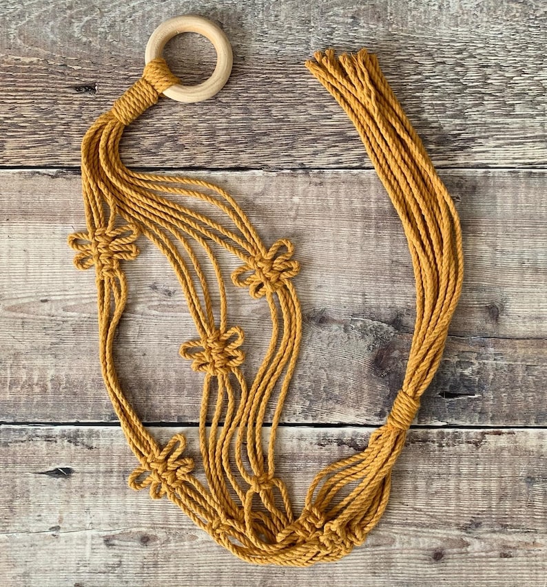Op de afbeelding: Een macram&eacute; plantenhanger met een houten ring aan de bovenkant en vijf geknoopte bloemmotieven. De hanger is gemaakt van naturel gekleurd koord.