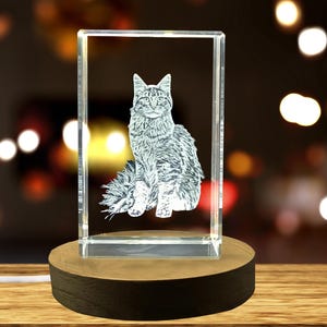 Peut inclure: Bloc de verre clair rectangulaire avec une image gravée d'un chat. Le chat est assis. Le bloc de verre est posé sur un socle rond en bois. L'arrière-plan est flou avec des lumières bokeh.