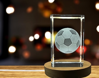Può includere: Un cubo di cristallo con un'immagine incisa in 3D di un pallone da calcio. Il cubo è su una base di legno ed è illuminato dall'interno.