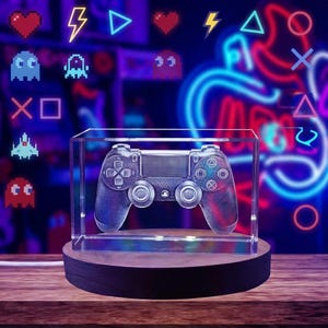May include: A clear rectangular glass block containing a laser-etched 3D image of a video game controller. The block rests on a circular dark wooden base. The controller features dual analog sticks, a directional pad, and standard action buttons.