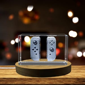 Op de afbeelding: Een heldere acryl display case met twee grijze gamecontrollers. De controllers worden weergegeven in een heldere doos, gemonteerd op een ronde houten basis. De achtergrond is vervaagd met bokeh-lichten.