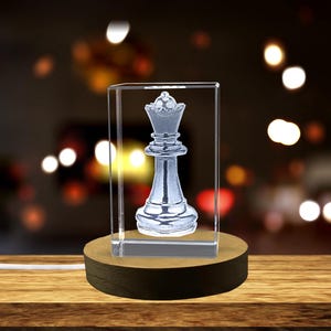 Könnte beinhalten: Ein klarer Kristallwürfel mit einem 3D-geätzten Design einer Schachdame. Der Würfel steht auf einem Holzsockel.