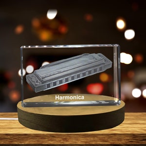 Op de afbeelding: Een heldere glazen kubus met een gegraveerde zilveren mondharmonica erin. De kubus staat op een houten voet. De tekst "Harmonica" is gegraveerd op de voet.
