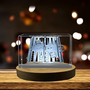 Könnte beinhalten: Ein Kristallwürfel mit einer 3D-gravierten Abbildung eines Backgammon-Bretts und Spielsteinen. Der Würfel steht auf einem Holzsockel mit einer Lichtquelle darunter.