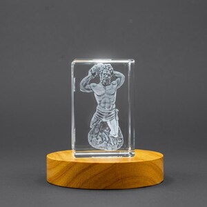 Op de afbeelding: Een helder rechthoekig glazen blok met een lasergeëtste driedimensionale figuur van een gespierde man die een steen boven zijn hoofd houdt, gemonteerd op een gladde, ronde, lichtgekleurde houten voet met een gepolijste afwerking.