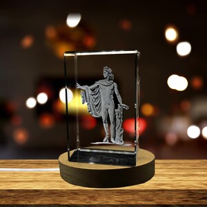 Può includere: Una scultura in vetro trasparente di una figura maschile classica, forse Apollo, all'interno di un blocco rettangolare. La scultura è grigia e poggia su una base di legno con una luce calda. Lo sfondo è sfocato con luci bokeh.
