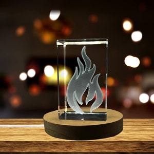 Op de afbeelding: Een heldere glazen kubus met een gegraveerd wit vlamontwerp staat op een houten basis. De kubus wordt van binnenuit verlicht, waardoor een warme gloed ontstaat.