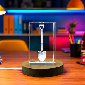 May include: A clear rectangular glass block featuring a laser-etched white graphic of a shovel in the center. The glass block is mounted on a circular dark wooden base that includes a white power cord extending from the side of the base.