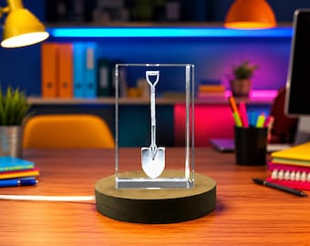 May include: A clear rectangular glass block featuring a laser-etched white graphic of a shovel in the center. The glass block is mounted on a circular dark wooden base that includes a white power cord extending from the side of the base.