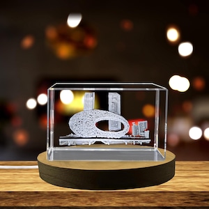 Może przedstawiać: Przezroczysty szklany sześcian z trójwymiarowym grawerem laserowym przedstawiającym nowoczesny budynek z zakrzywionym dachem. Sześcian stoi na drewnianej podstawie.