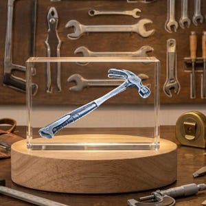 May include: A silver hammer with a black grip is encased in a clear acrylic block, displayed on a wooden base. The background features various tools, including wrenches, saws, and a tape measure, suggesting a workshop or tool-themed setting.