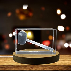 Peut inclure: Un cube en verre transparent avec une image gravée en 3D d'un maillet noir et argent. Le cube est posé sur une base en bois.