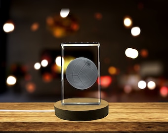 Può includere: Una palla da pallavolo grigia racchiusa in un cubo di vetro trasparente su una base di legno. Il cubo è illuminato dal basso, creando un effetto 3D.